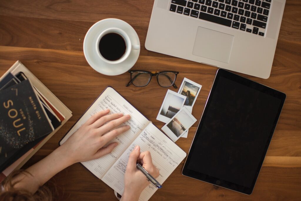 hands writing in planner with coffee, photos, and laptop in view.